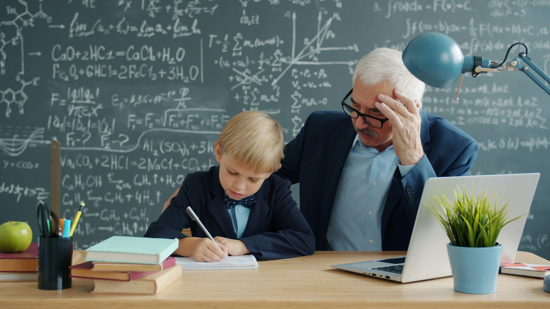 Een klein jongetje die iets opschrijft in een kladboek met een professor naast zich en een krijtbord op de achtergrond