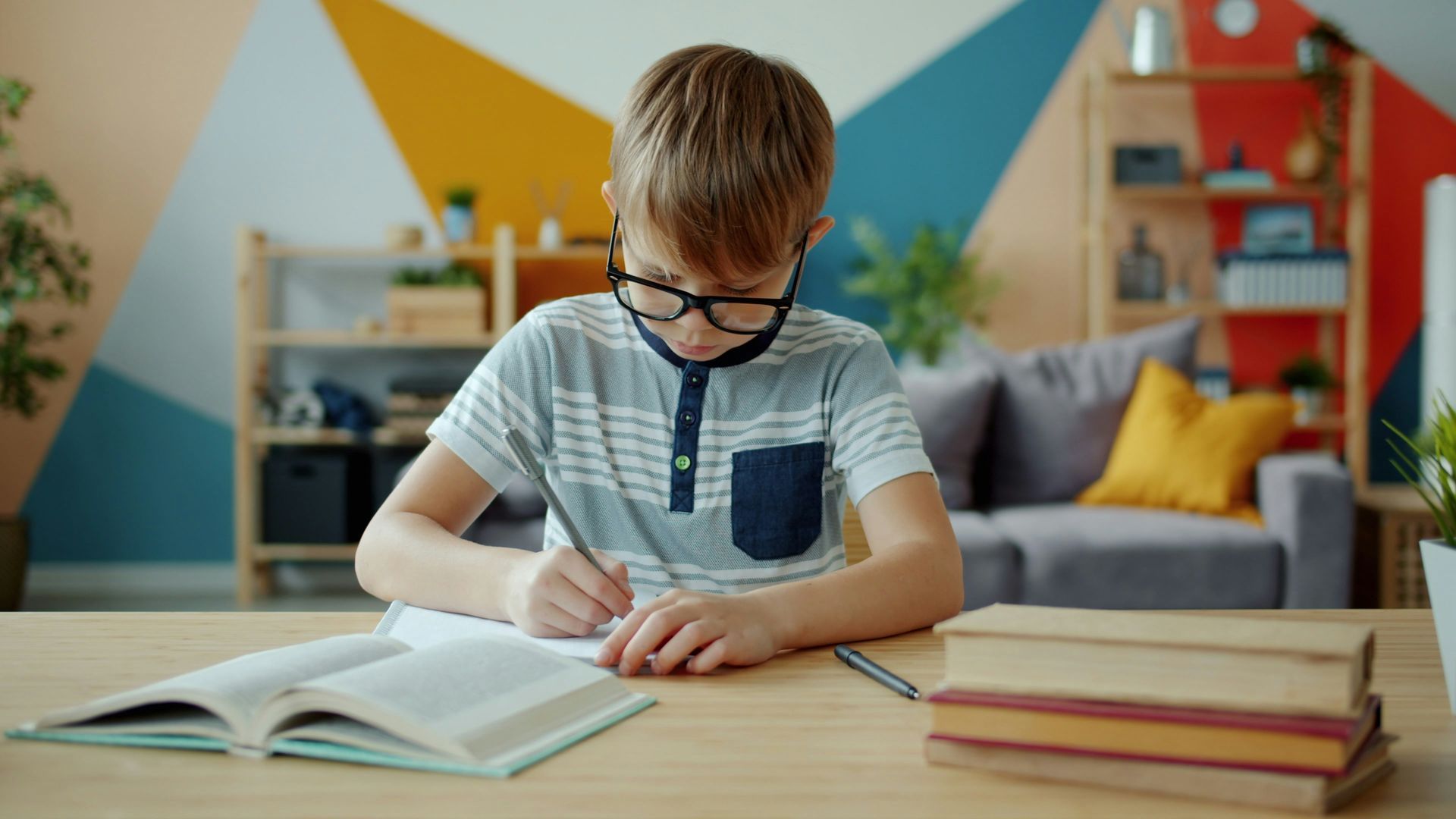 Jongetje met een bril zit aan een bureau met boeken om hem heen en schrijft in een schrift