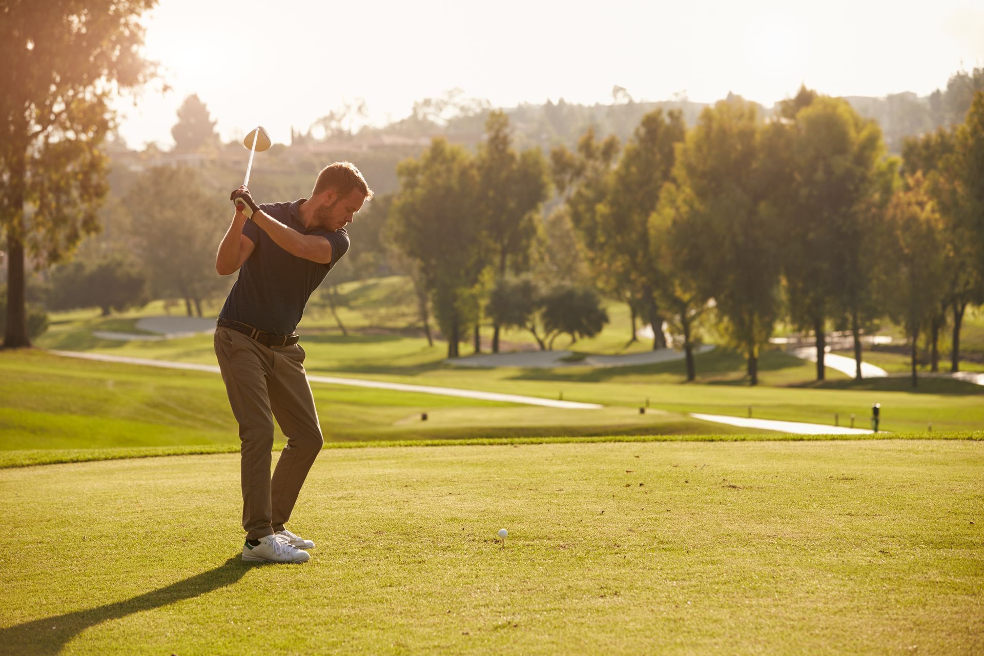 A golfer prepares to swing on a sunny day at a lush golf course, surrounded by trees.