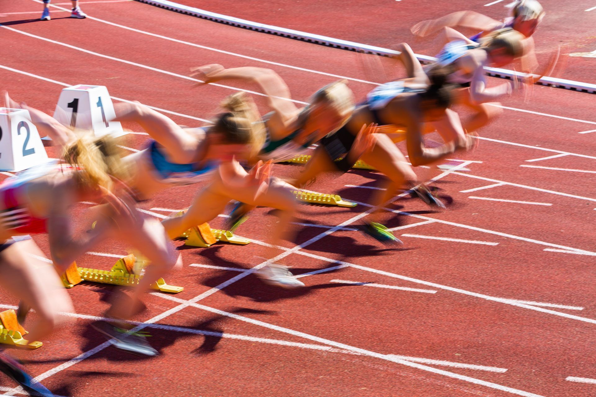 Runners take off from starting blocks on a track, blurred by speed in a race.