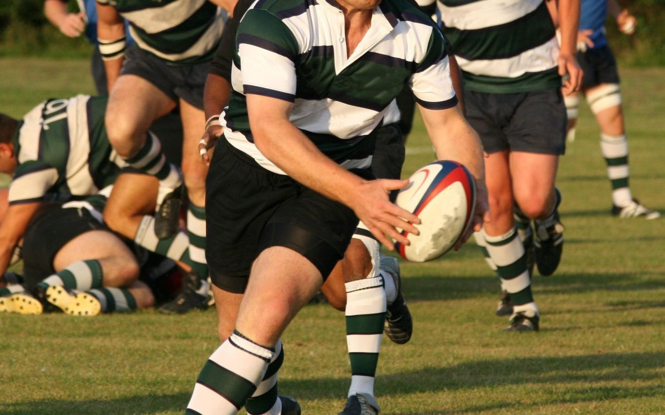 A rugby player in a striped jersey runs with the ball mid-game on a grassy field, while teammates and opponents follow behind.