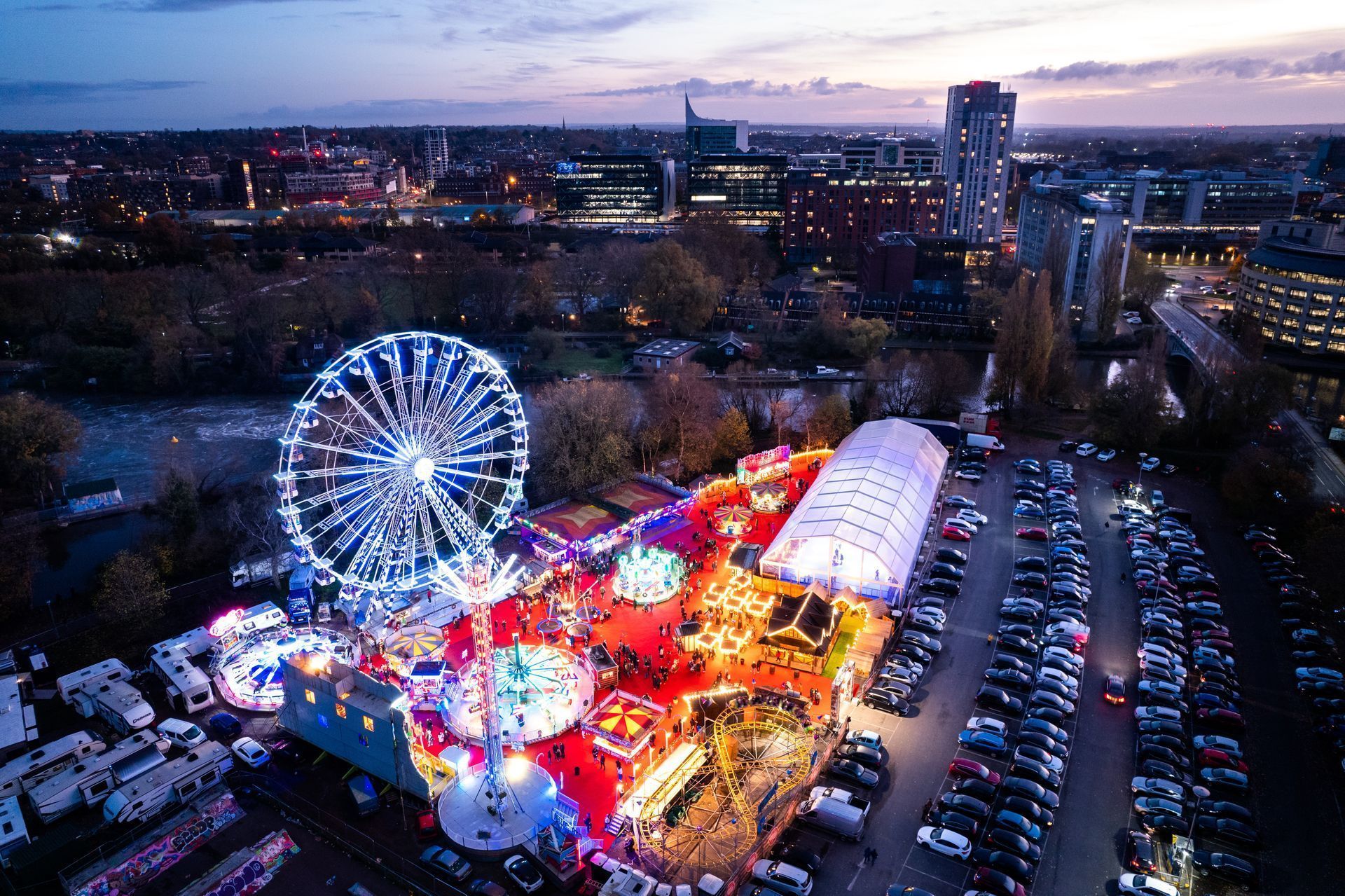 Aerial view of a brightly lit amusement park at dusk with a ferris wheel, various rides, tents, and parked cars. City buildings and a sky with clouds are visible in the background.