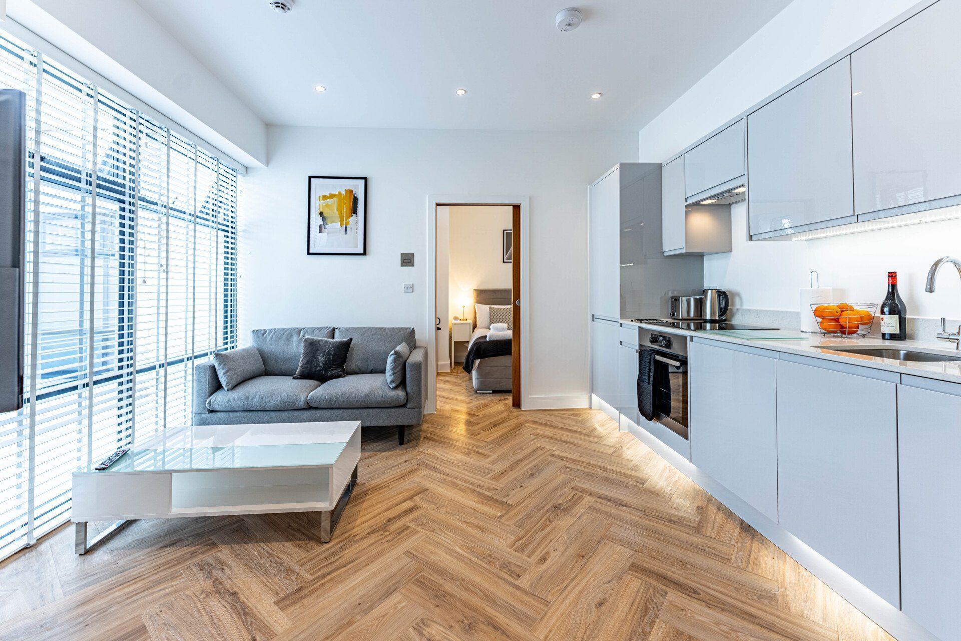 Modern open-plan living area in Wellington House with a grey sofa, glass coffee table, and a fully equipped kitchen. A door leads to a bedroom in the background. Wooden herringbone floors and large windows provide natural light.