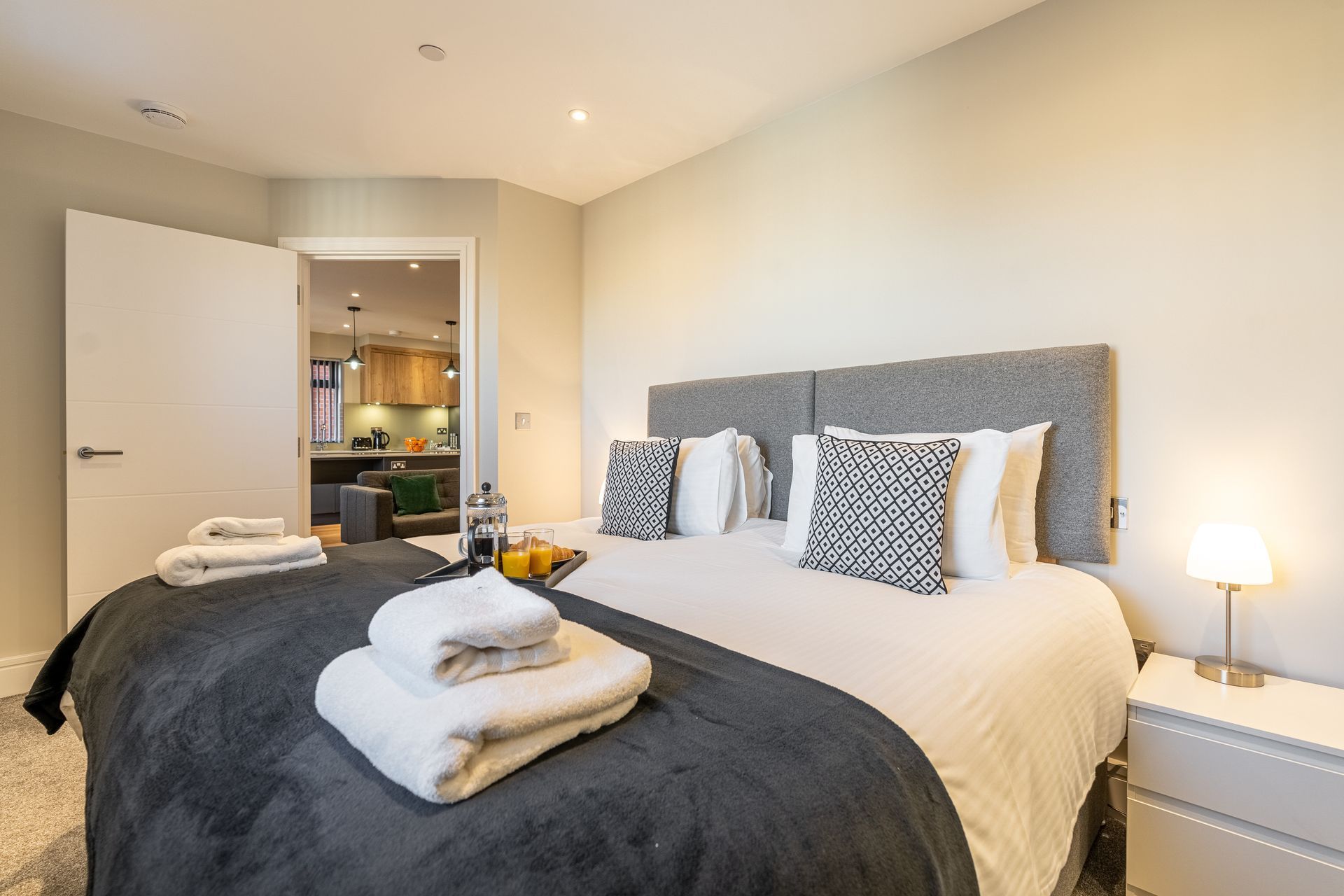 Modern bedroom with twin beds, gray headboards, folded towels, and an open door to a kitchen.