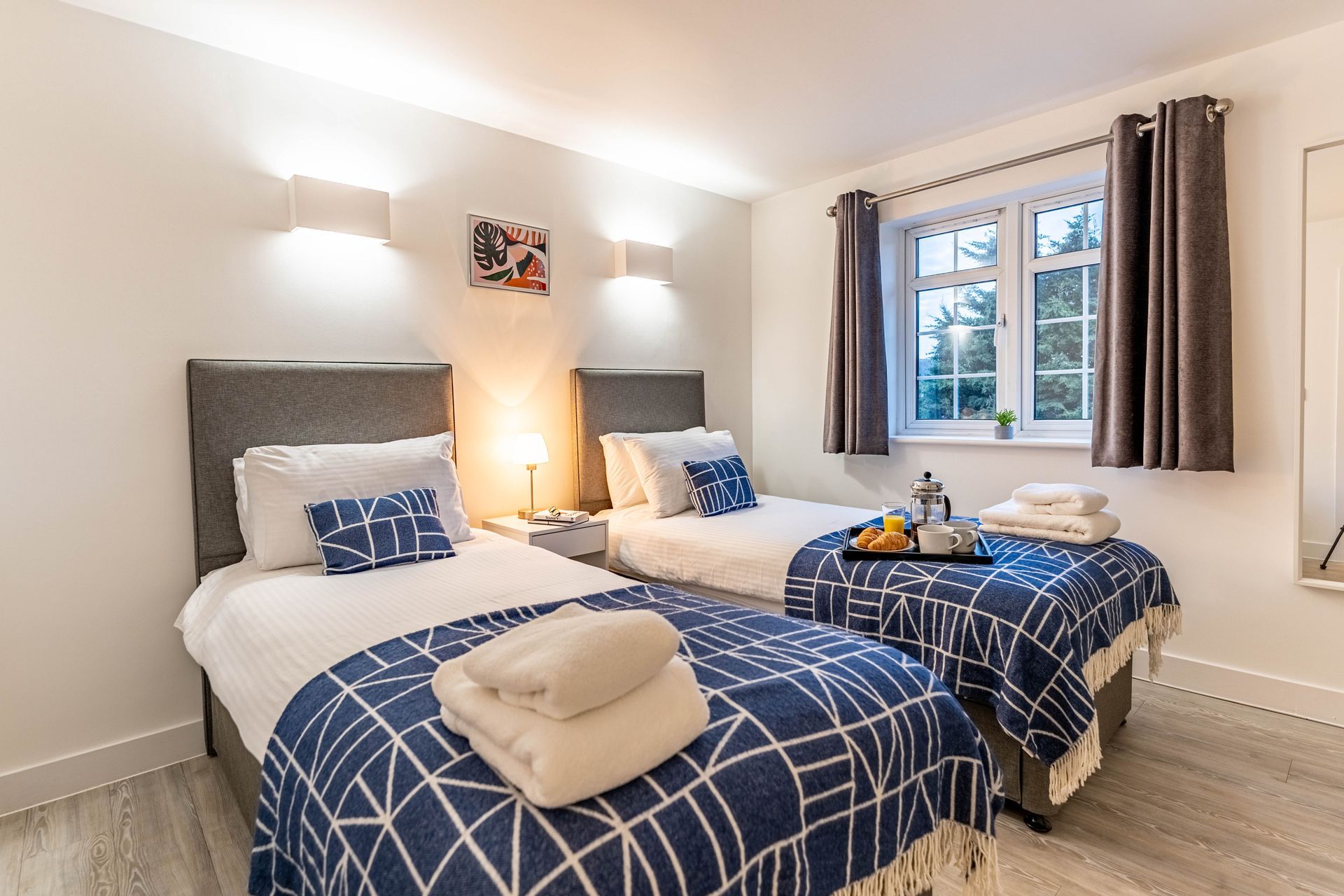 A hotel room with two twin beds featuring white bedding, blue patterned throws, folded towels, and a window with curtains.