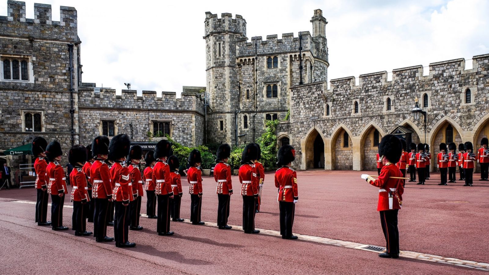 A formation of soldiers in red uniforms and black hats stands in front of a historic stone building with arches and turrets.