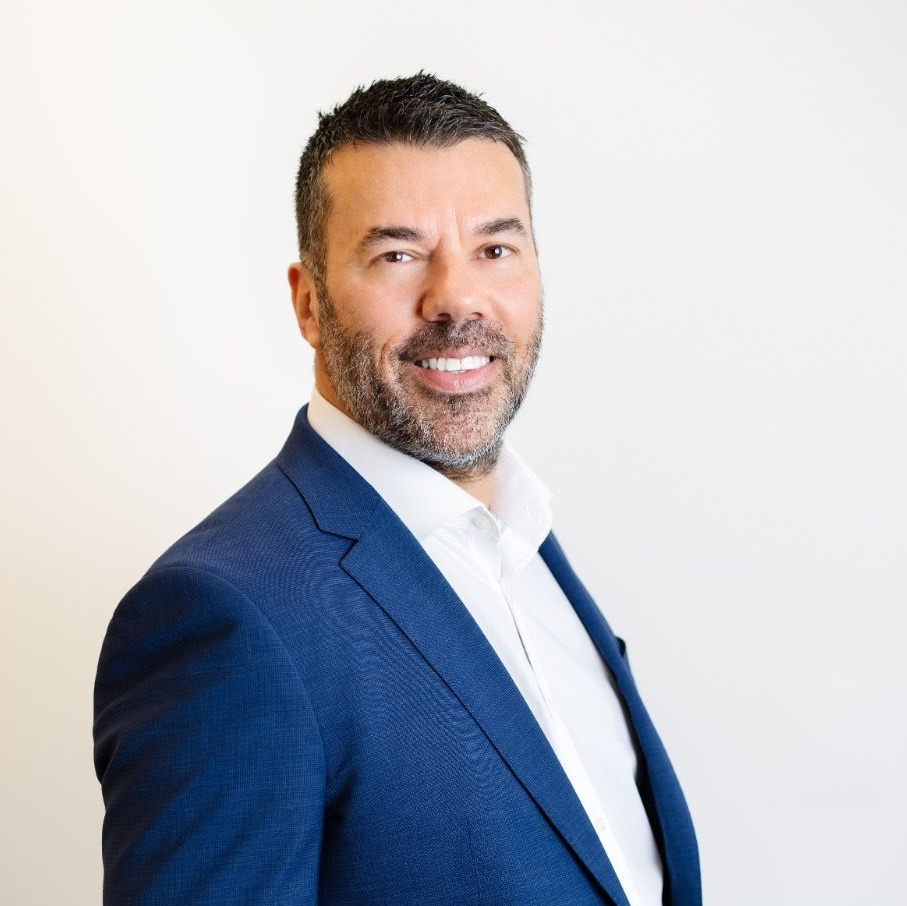 A professional headshot of a smiling man with a short beard, wearing a blue blazer and white shirt against a white wall.