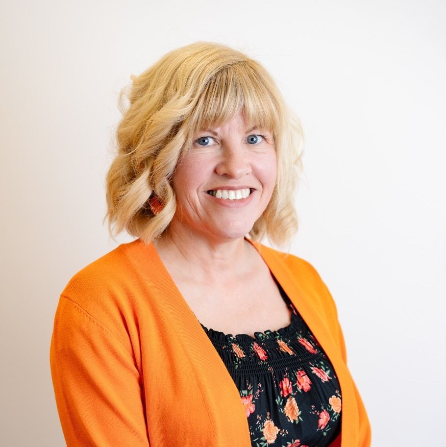 A smiling woman with blonde hair, wearing a black floral top and an orange cardigan against a white background.