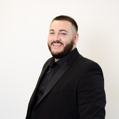 A person with a beard, wearing a black suit and shirt, smiles against a plain white background.