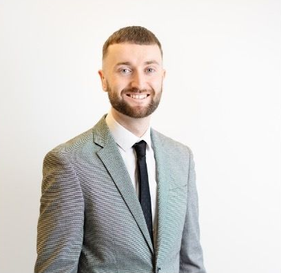 A smiling professional wearing a gray textured blazer, white dress shirt, and black necktie against a white background.