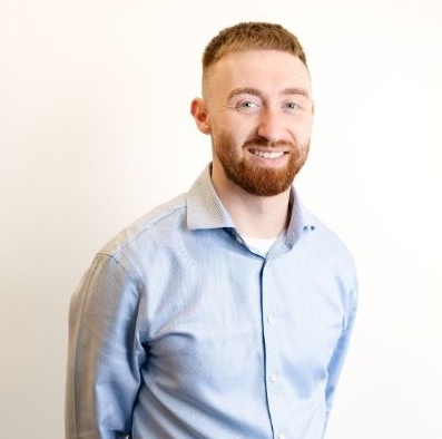A man with a short beard smiling, wearing a light blue collared shirt against a plain white background.