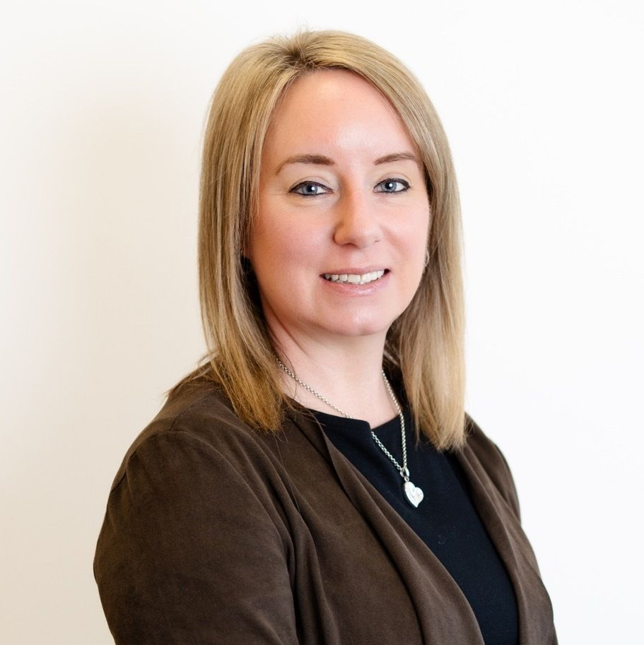 A professional headshot of a person with shoulder-length blonde hair, wearing a brown jacket over a black shirt.
