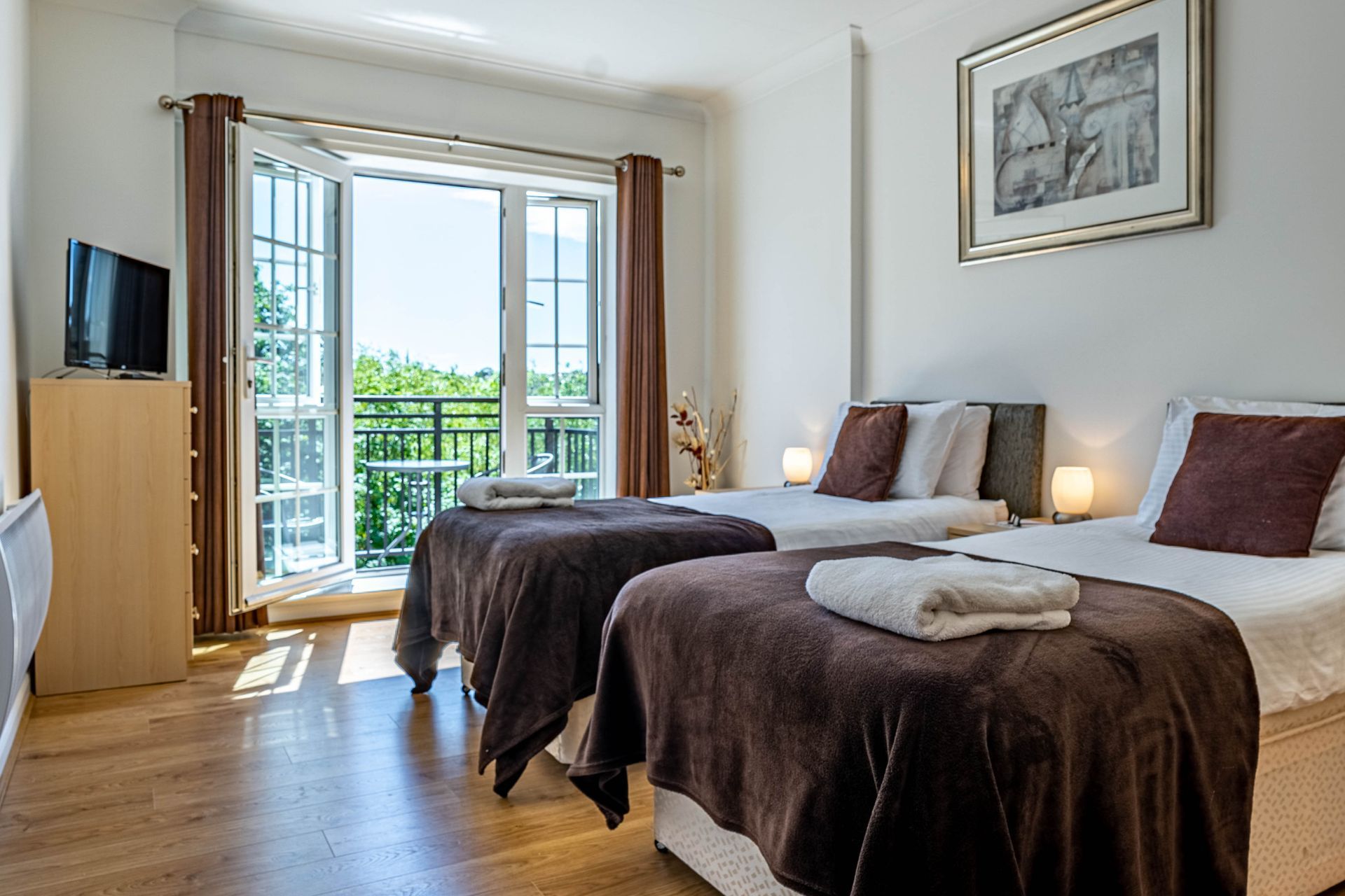 A hotel room featuring two twin beds with brown blankets, a TV on a stand, and a balcony with open glass doors.