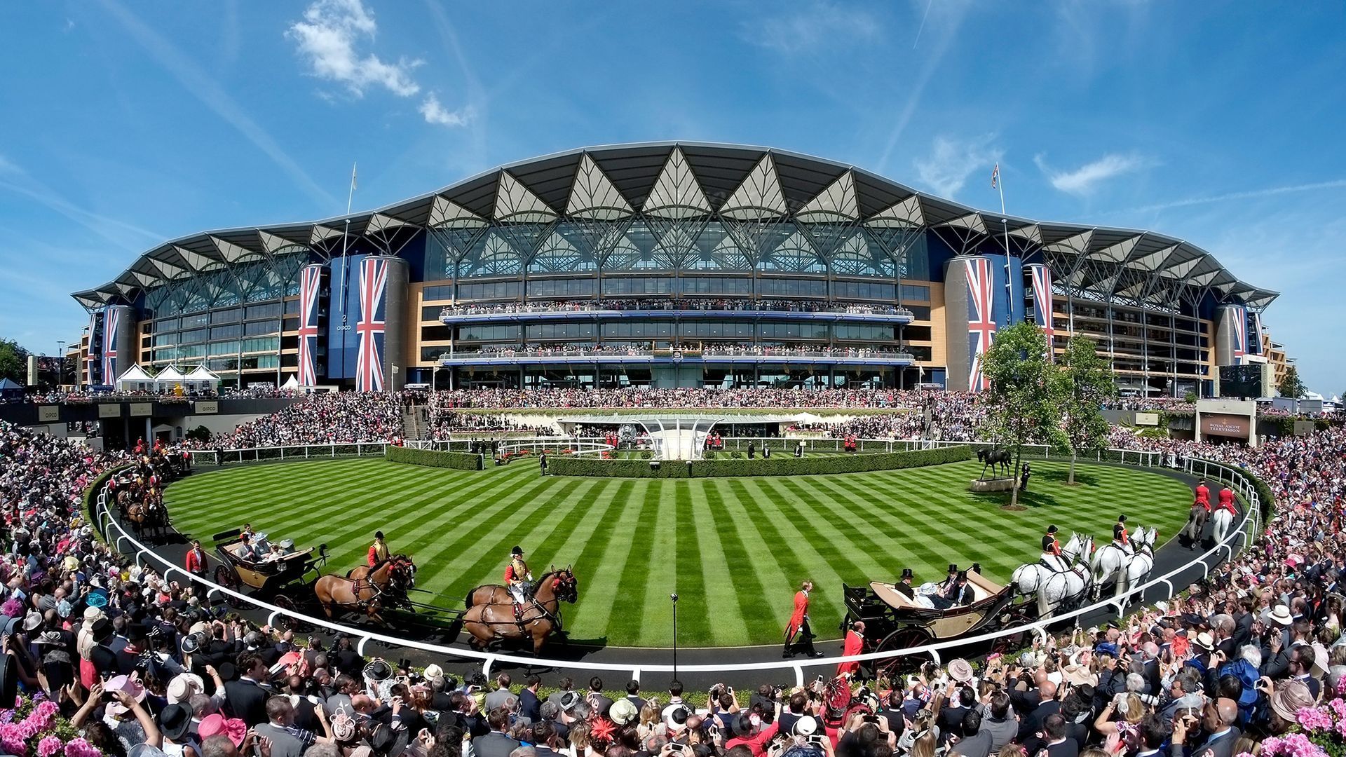A large crowd gathers around a racetrack at a stadium with Union Jack decorations. Horse-drawn carriages and well-dressed individuals are visible on the track. The sky is clear and blue.