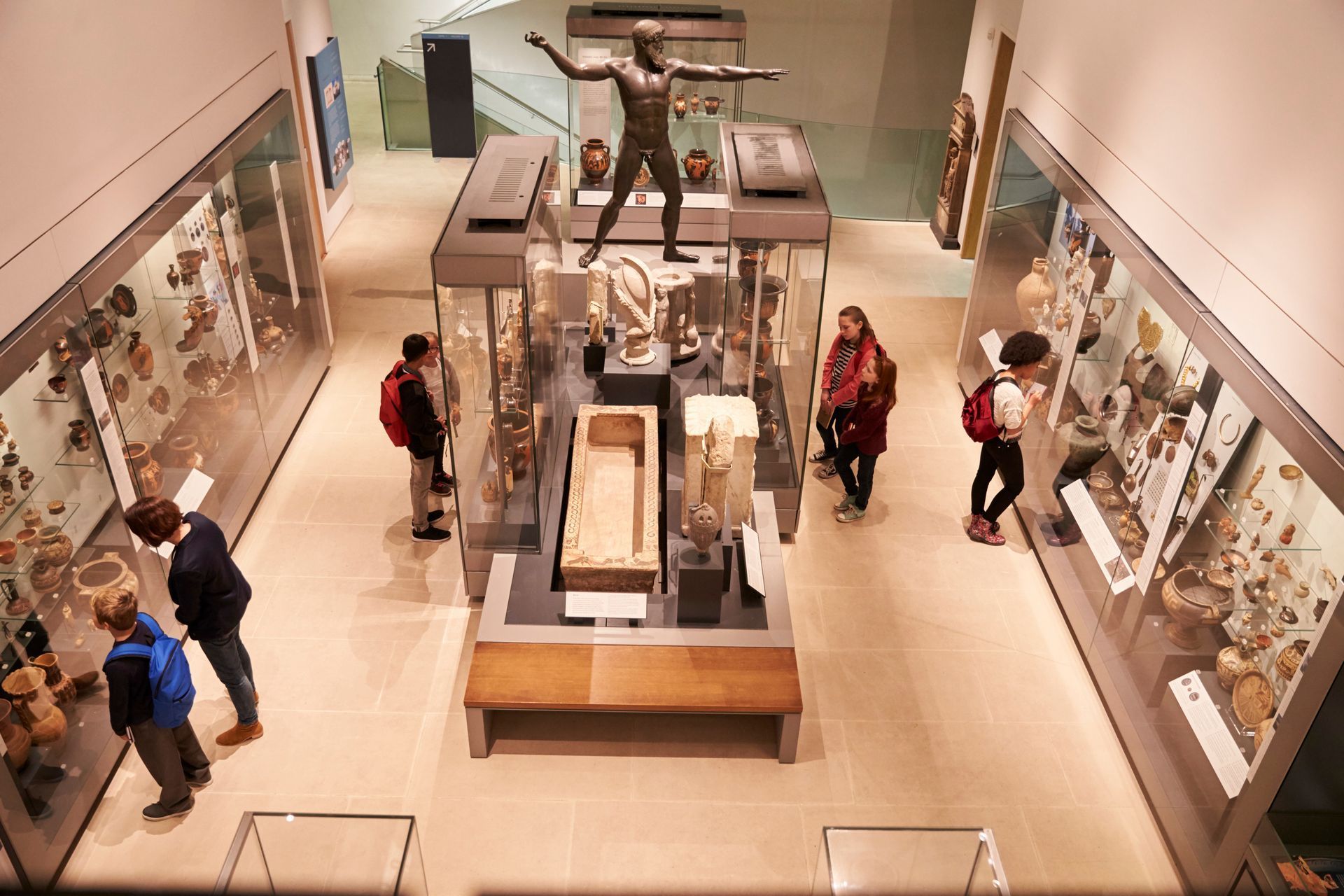 Visitors explore a museum gallery featuring antiquities, including a central bronze statue and glass display cases.