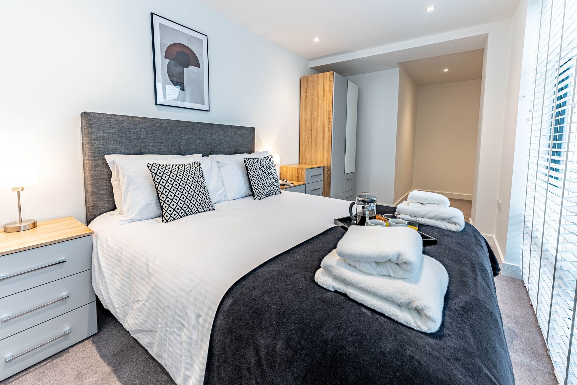Modern bedroom with a neatly made bed, folded towels, and a tray with cups on the blanket.