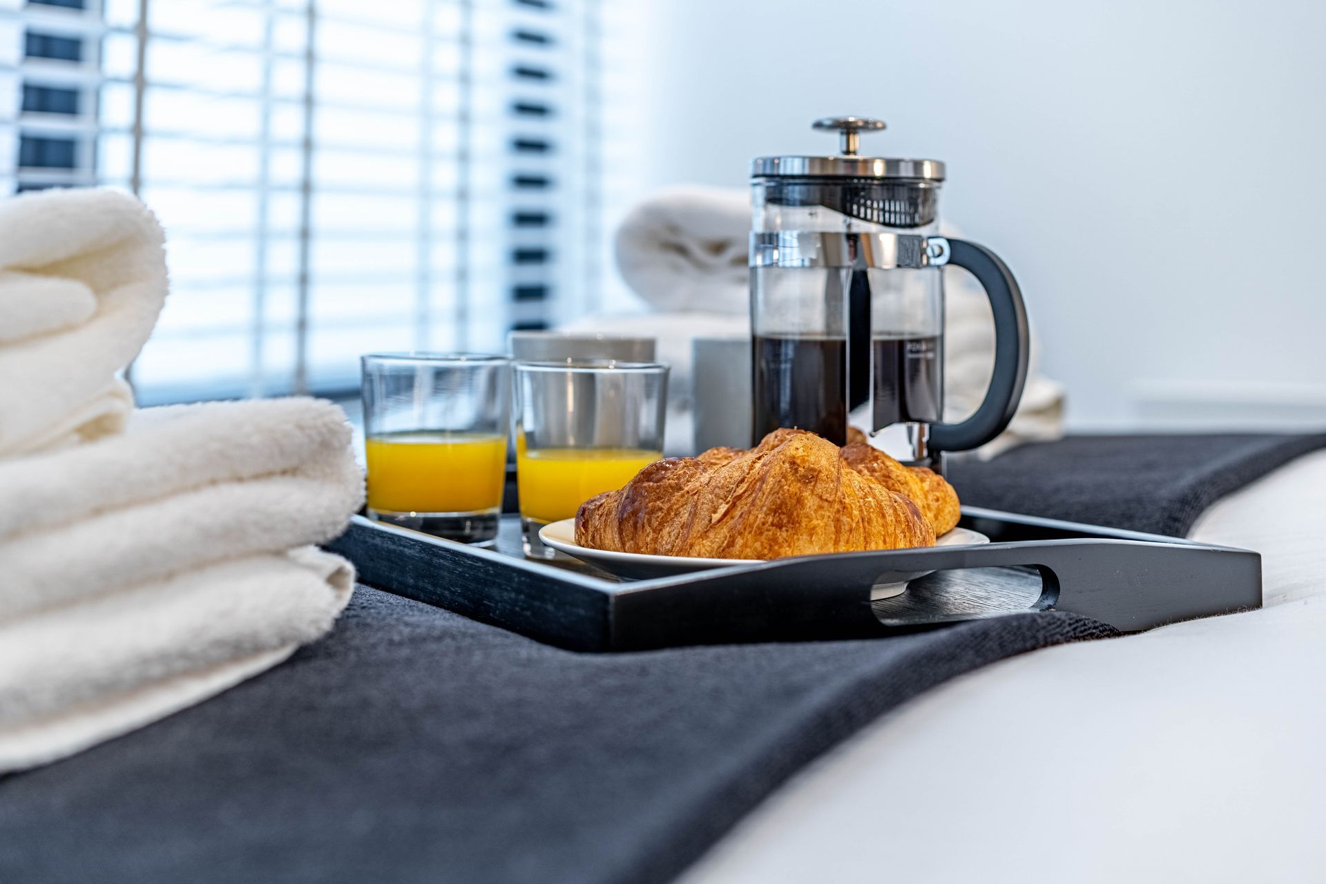 A breakfast tray with a croissant, two glasses of orange juice, and a French press coffee maker on a bed with white towels beside it.