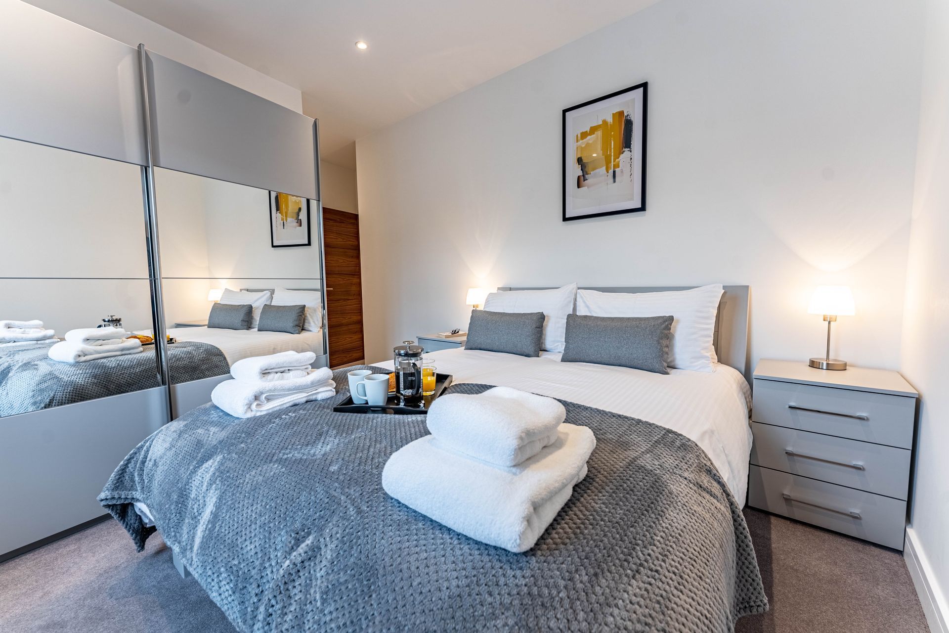 A modern bedroom with a bed topped with white folded towels, a grey throw blanket, and a small tray with drinks.