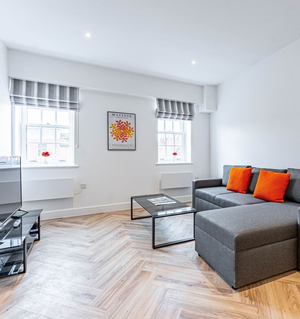 A bright living room with herringbone wood floors, a grey sectional sofa with orange cushions, and two windowed walls.