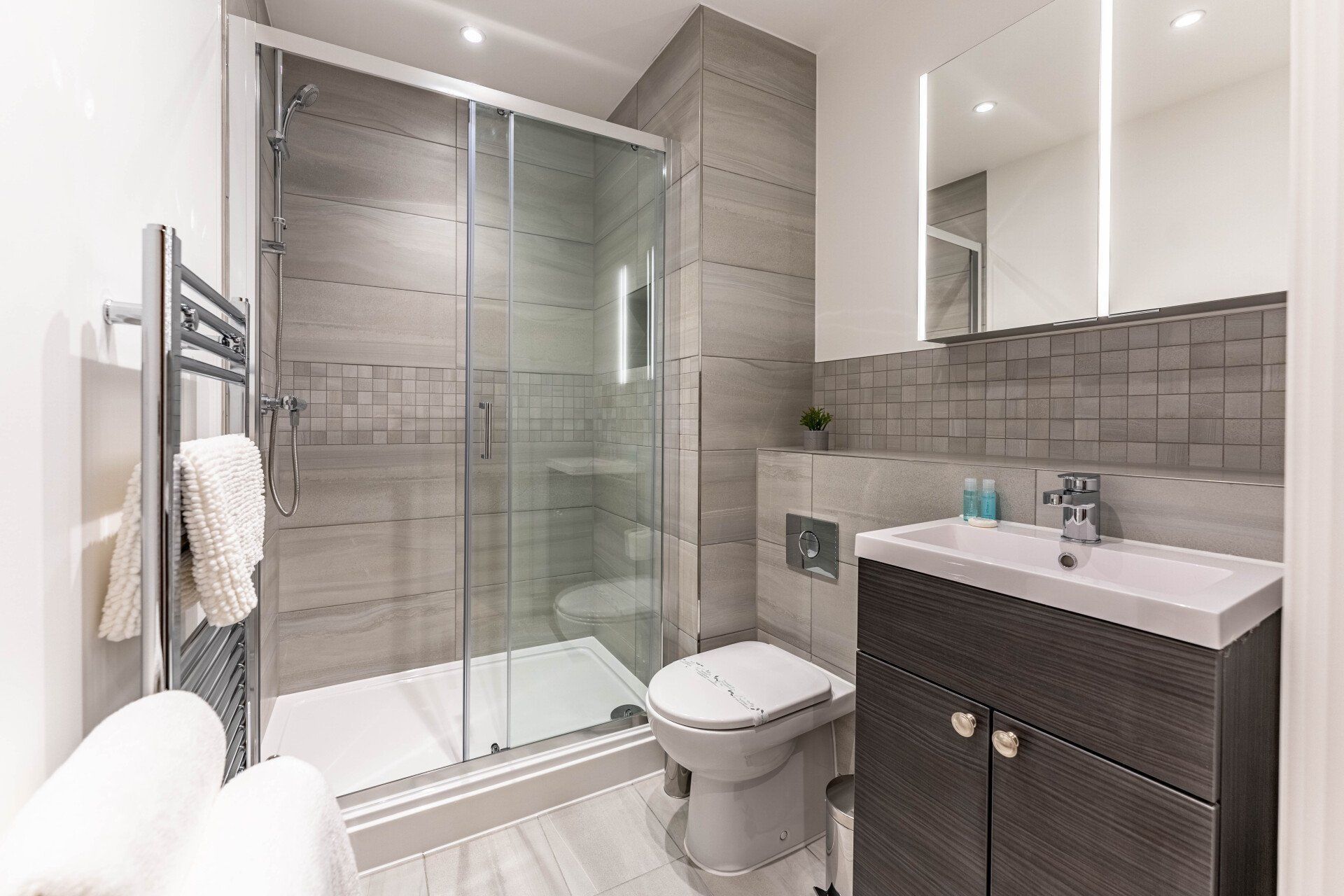 Serviced Apartment in Reading - Modern bathroom at Wellington House Reading featuring a glass-enclosed shower, toilet, dark wood vanity with a sink, and a mirrored cabinet. Tiled walls and floors in light gray tones complement the overall design.