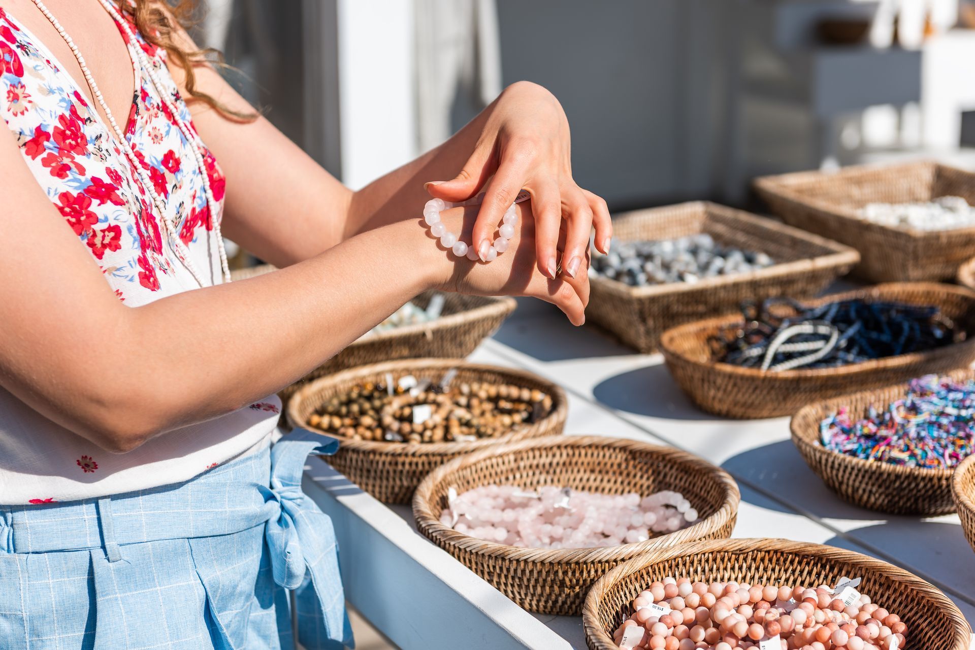 A person tries on a light-colored beaded bracelet at an outdoor market filled with baskets of gemstone jewelry.
