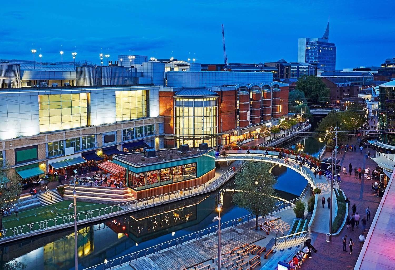 An evening view of a canal-side shopping and dining area in Reading, UK, with illuminated buildings and a pedestrian bridge.