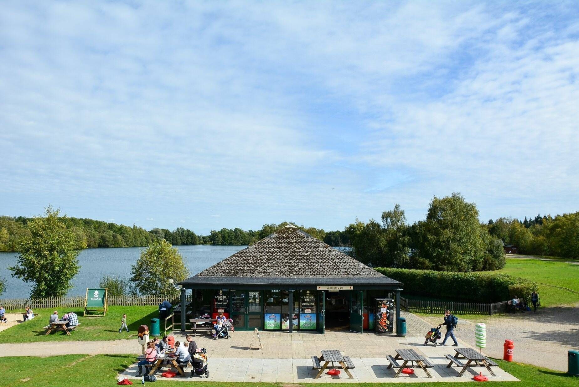 People sitting at picnic tables near a small building by a lake; green trees and a partly cloudy sky surround the area.