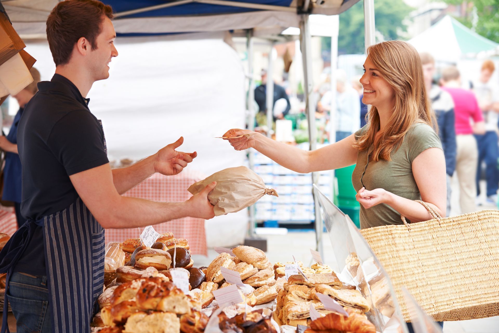 A customer receiving a paper bag from a vendor at a bakery stand in an outdoor market.