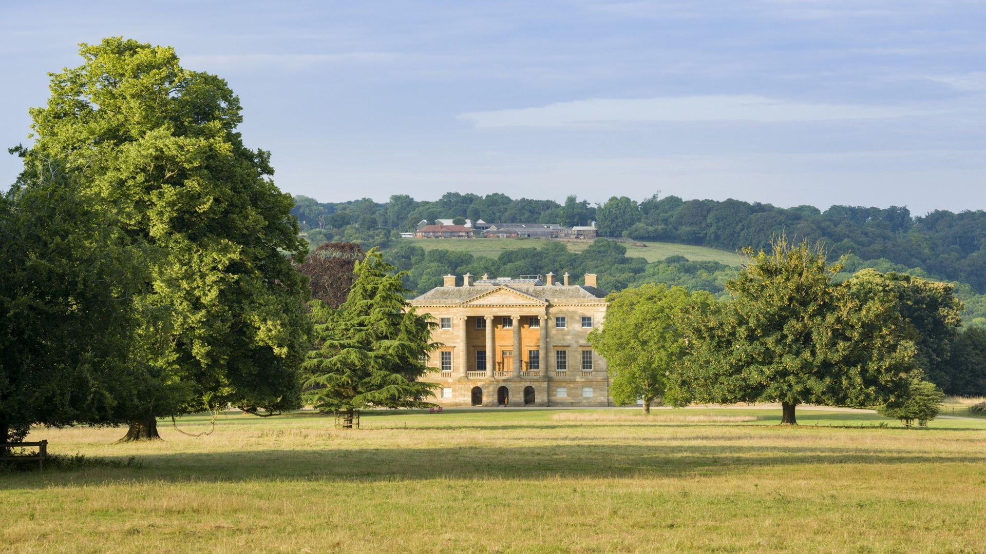 A large, stately mansion with classical columns is set amidst a vast, green lawn and surrounded by trees, with hills in the background under a partly cloudy sky.