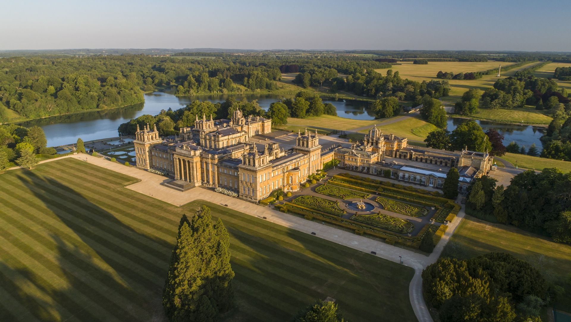 Aerial view of a large, historic palace surrounded by manicured gardens, expansive lawns, and a lake. The surrounding area is lush and green with trees and open fields.
