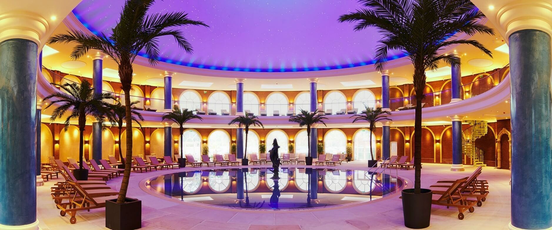 A luxurious indoor pool area with palm trees, lounge chairs, and a dome ceiling illuminated in blue and purple lights.