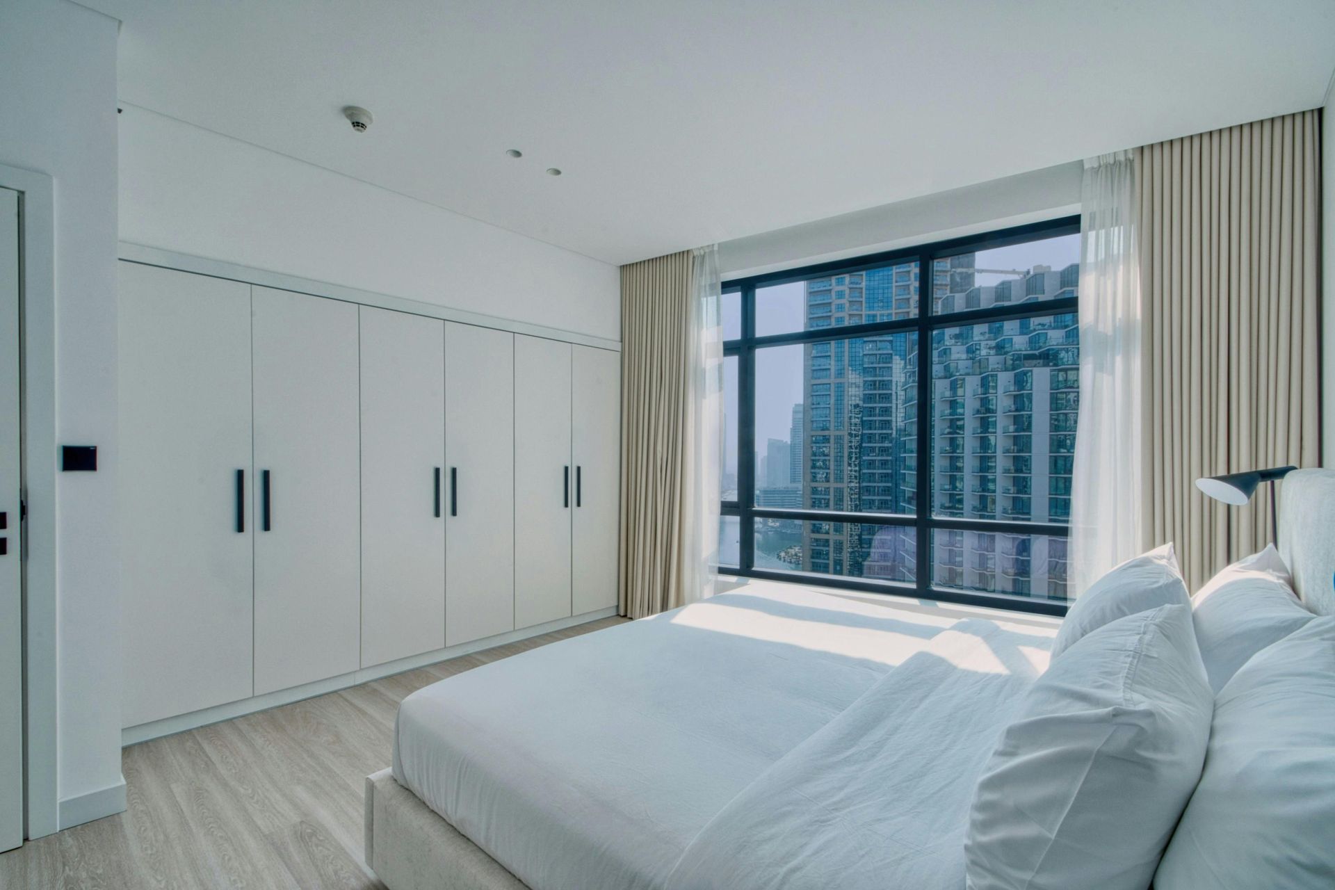 A modern, minimalist bedroom with white walls, floor-to-ceiling closets, and a large window overlooking a city skyline.