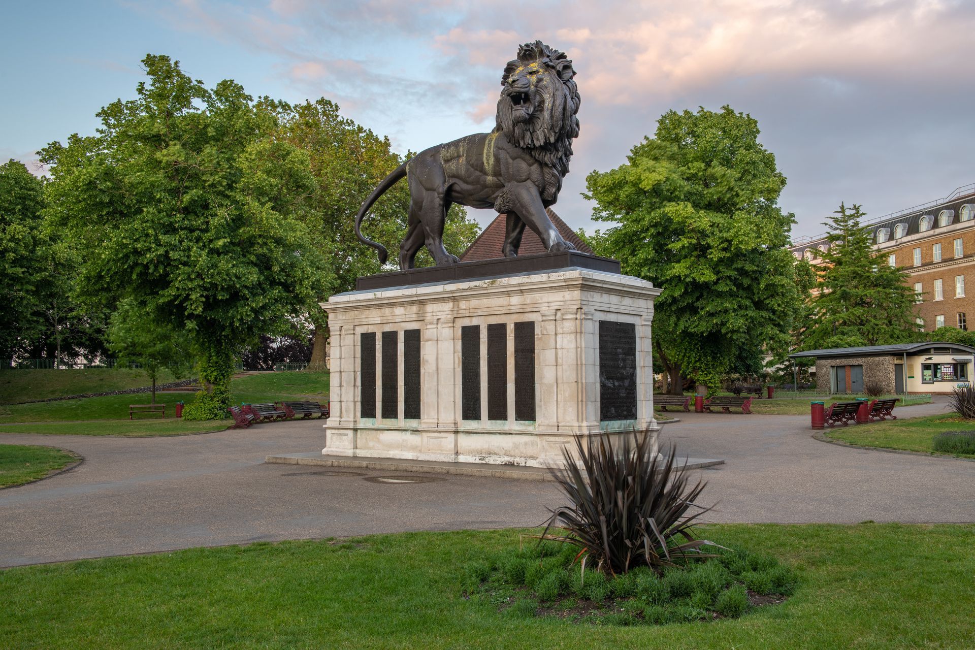 A bronze lion statue stands atop a stone memorial plinth in a lush green park during late afternoon.