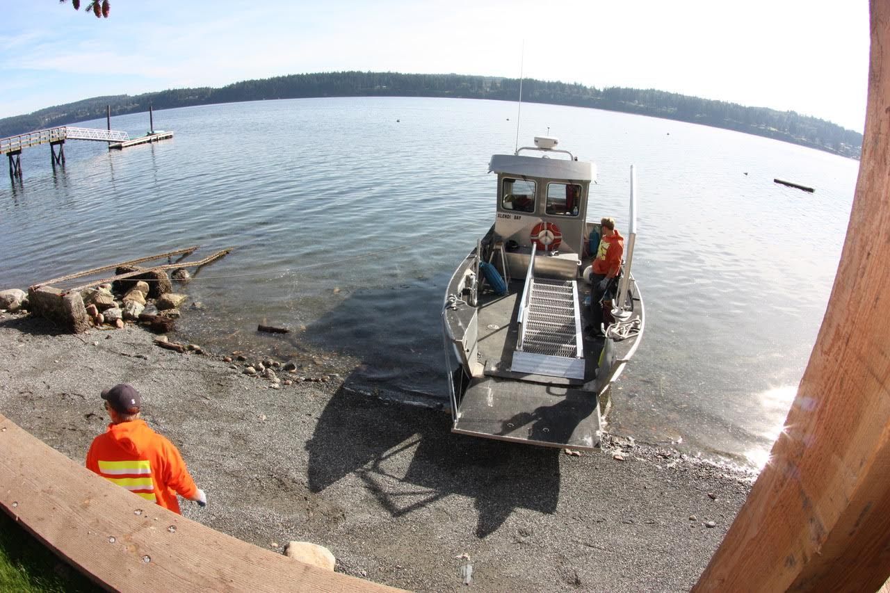 Small boat at a rocky lakeshore with two people in orange safety vests and a dock nearby