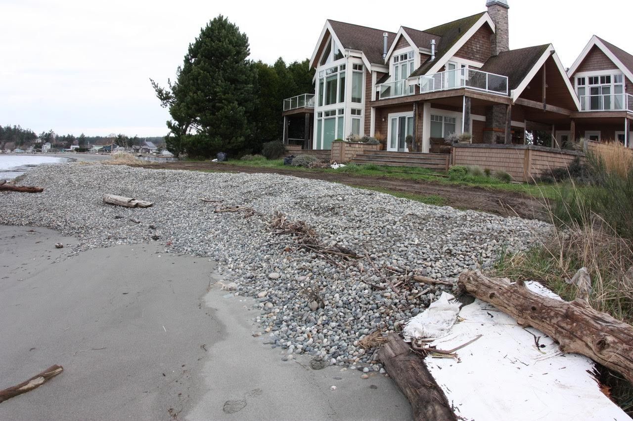 Pebble-strewn beach beside a large house with a seawall and overcast sky