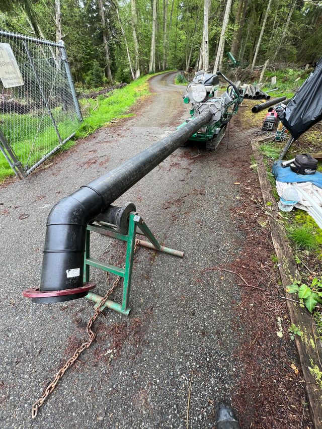 Black pipe laid along a gravel path in a wooded area, with metal supports and debris nearby.