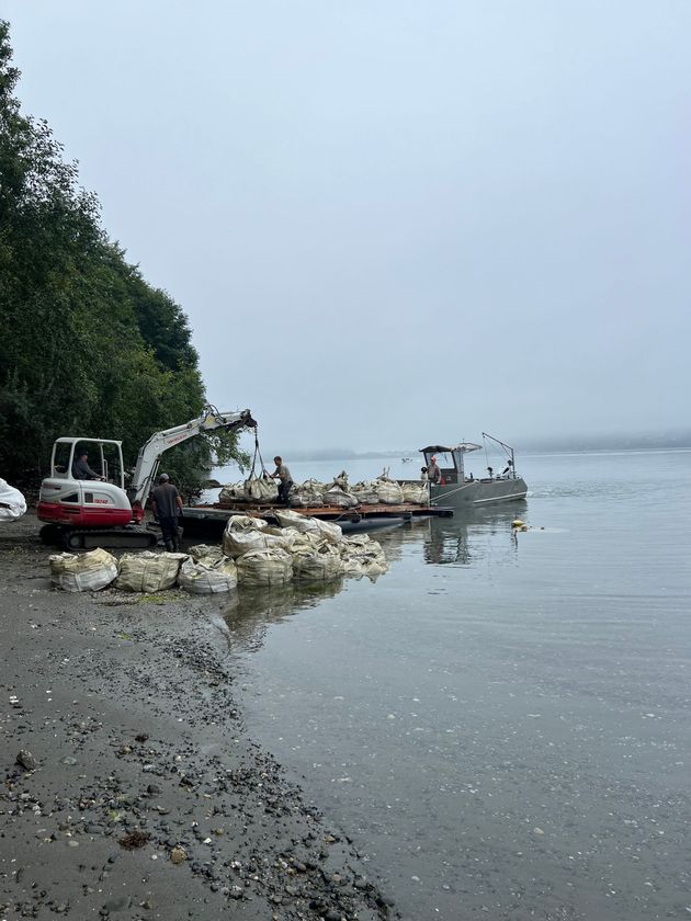 Beached boats and excavator on a rocky shoreline beside calm water under an overcast sky
