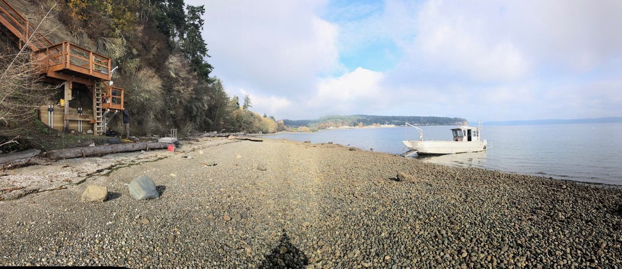 Rocky beach with a small white boat near shore, calm blue water, and a cliffside house on the left.