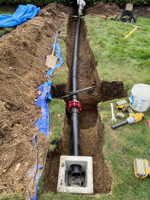 Trench with exposed black pipe and red valve, digging tools, and blue tarp beside a grassy yard.