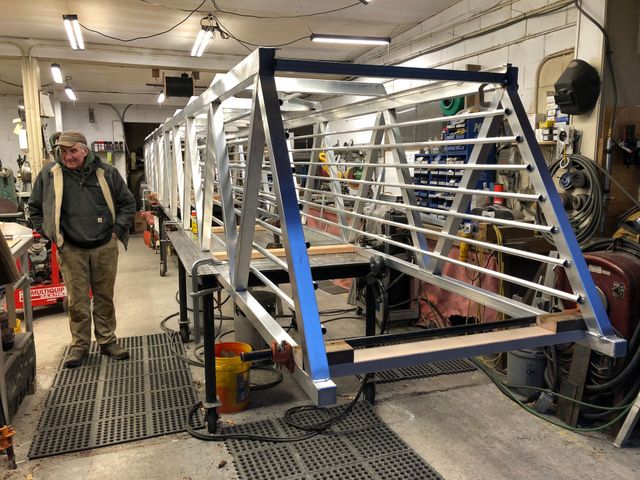 Person in a workshop beside a large metal frame and machinery, with tools and racks in the background