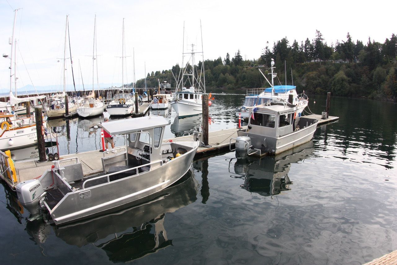 Small boats docked at a marina on calm water, with forested shoreline in the background