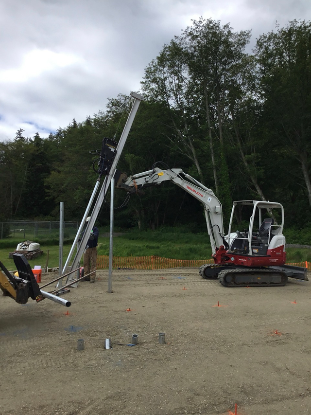 Excavator and construction equipment lifting a long metal pole on a gravel site near trees