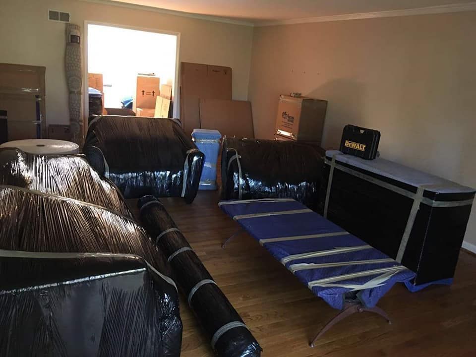 Room packed for moving: furniture wrapped in black plastic, boxes, rolled rugs, a cot.
