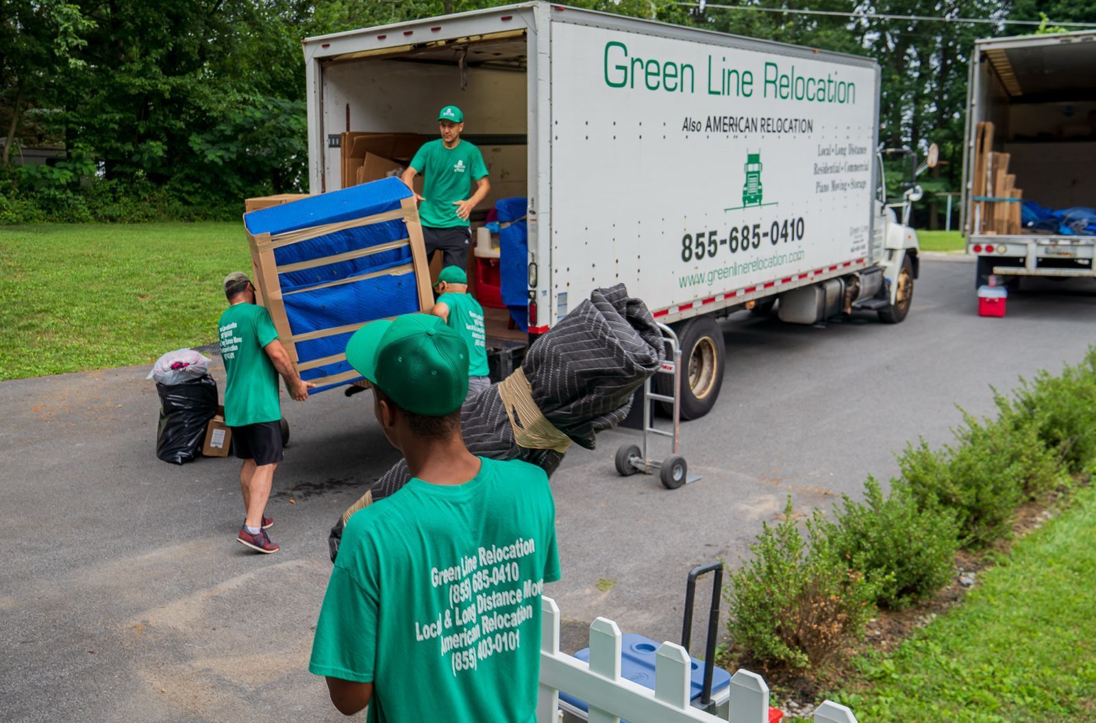 Movers in green shirts loading boxes into a Green Line Relocation truck on a paved driveway near a grassy area.