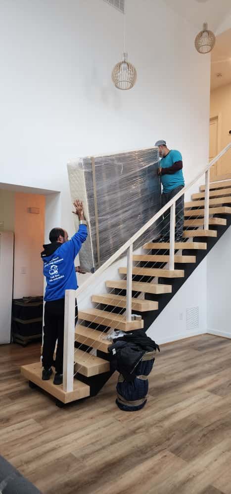 Two movers carrying a large wrapped object up wooden stairs. One is above, one below.