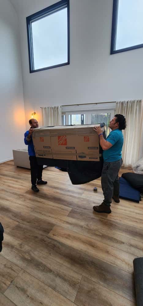 Two people carrying a large box into a light-filled room with wooden floors.