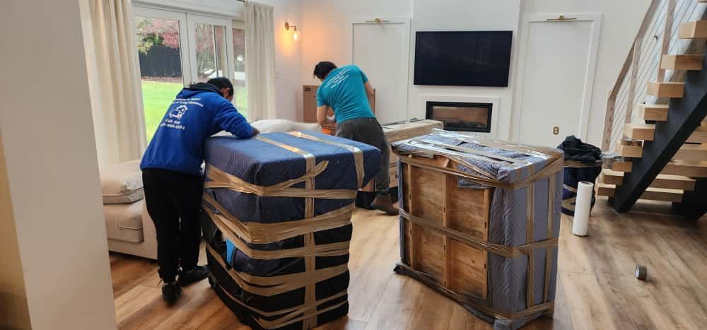 Movers carrying wrapped furniture in a living room with stairs and a window in the background.
