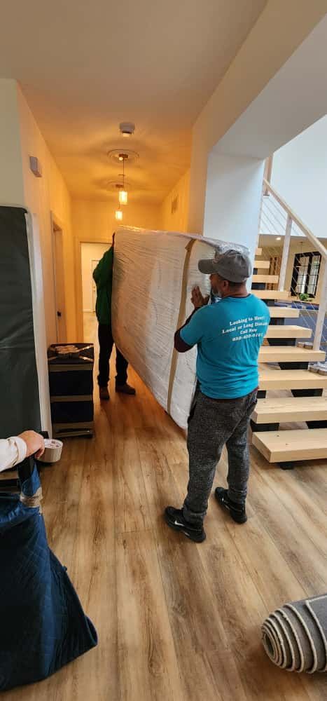 Two movers carrying a large, wrapped object down a hallway; a third person is partially visible.