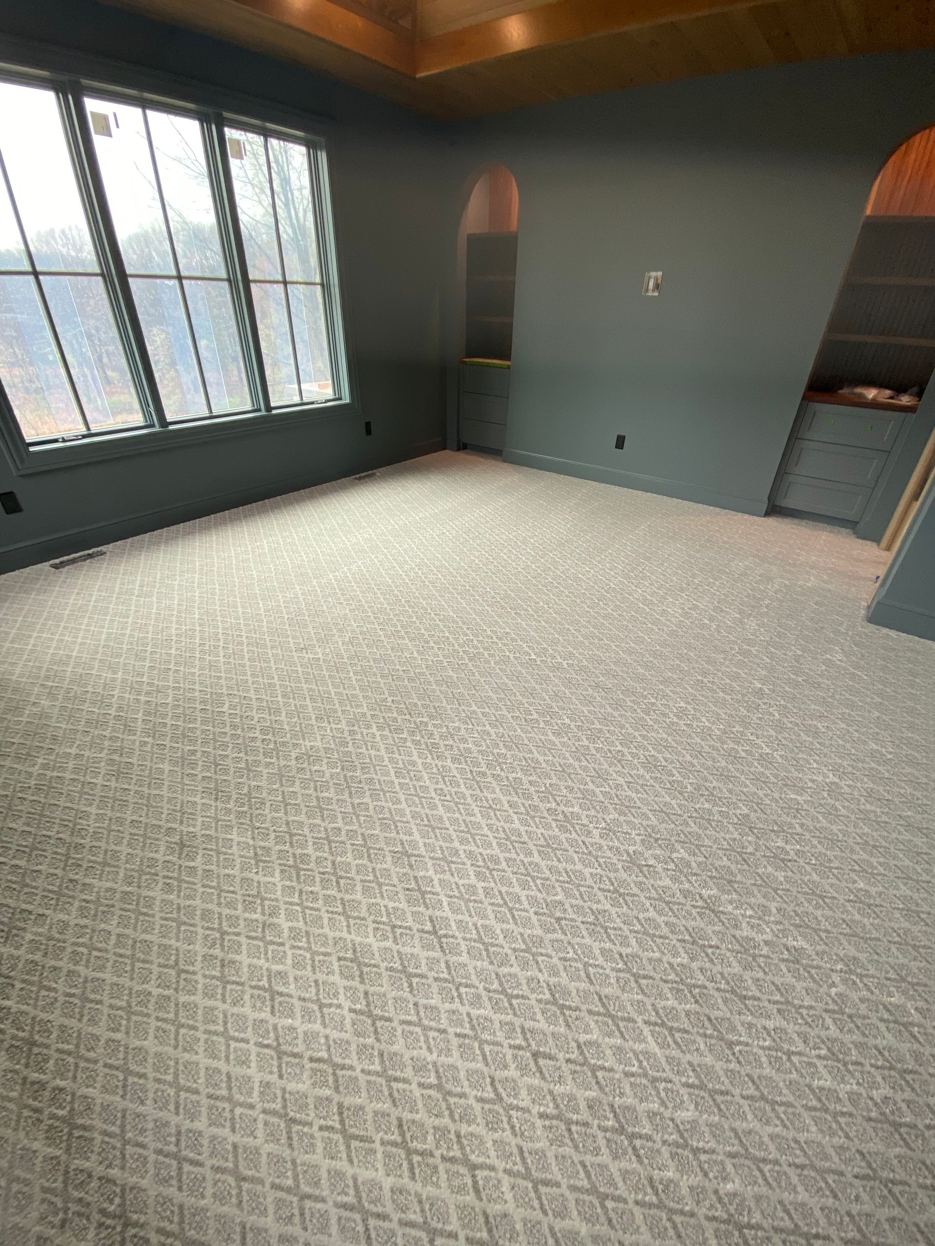 Empty office room with patterned carpet, blue-gray walls, built-in shelves, and a large window.