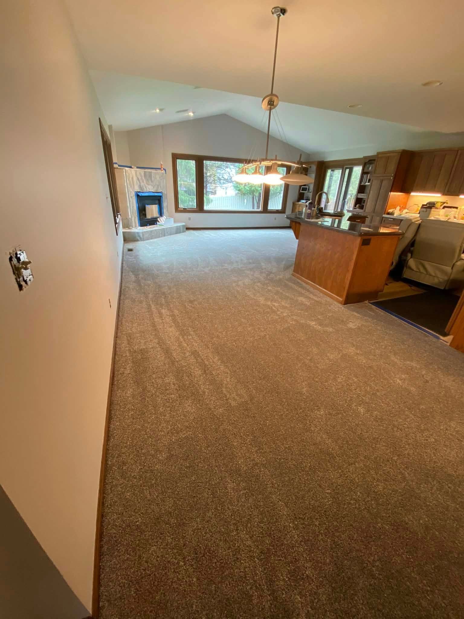 Spacious carpeted living room with vaulted ceiling, large window, wood cabinets, and kitchen counter area.